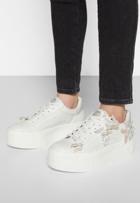 Bílé platformové sneakers boty z texturované kůže, s šněrováním a dekorativními motýlkovými akcenty v metalických tónech po stranách.