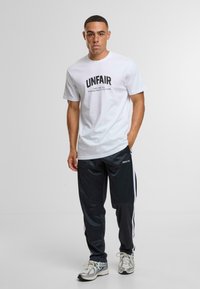 Fehér pamut póló, fekete "UNFAIR" felirattal, fekete, rugalmas derékú nadrággal párosítva, amelyen fehér oldalsávok vannak, valamint textúrázott sportcipőkkel.