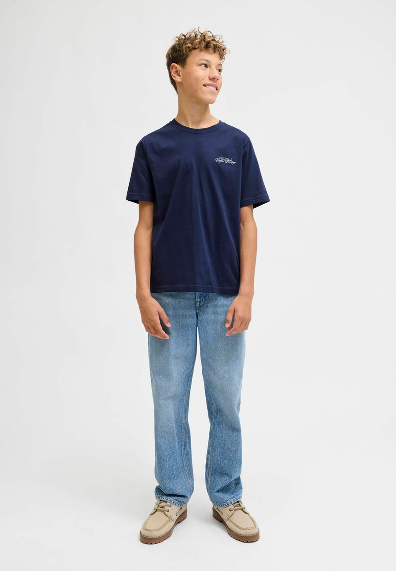 Tamsiai mėlynas medvilninis marškinėliai su apvalia iškirpte, derinami su šviesiai mėlynomis tiesiomis kelnėmis ir smėlio spalvos bateliais su raišteliais; minimalus dekoravimas ir minkšta tekstūra.