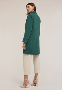 Cappotto verde in misto lana con colletto a revers, taglio dritto e due tasche laterali. Abbinato a pantaloni crema e scarpe con tacco nude.