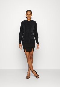 Blusa preta brilhante de mangas longas com gola alta, combinada com uma saia preta justa com duas fendas laterais e sandálias com tiras no tornozelo.