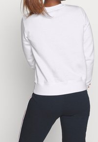 Kvinna med långt hår som bär en vit sweatshirt och marinblå byxor med vita och röda stadsremsor, vänd bort mot en enkel bakgrund.