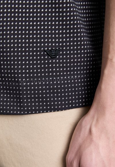 Dark patterned shirt featuring small white triangles, with a textured surface and an embroidered black logo on the lower left side.