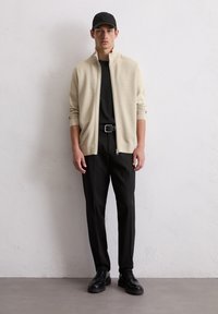 Lichtbeige ritsjack over een zwart t-shirt, gecombineerd met zwarte broeken en schoenen, gepresenteerd tegen een neutrale achtergrond.