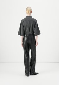 Zwarte denim blouse met wijde mouwen, gezien van achteren, gecombineerd met hoge zwarte neplederen broeken en haklaarzen.