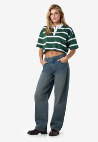 Kortermet, grønn polo med hvite striper, kombinert med løstsittende blå denimjeans og svarte ankelstøvler.