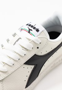 Sneaker bianchi in pelle con un design a swoosh nero, superficie texture, lacci bianchi e un'etichetta con logo che presenta accenti verdi e rossi.