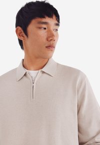 Beige, leichter Strickpullover mit einem Reißverschlusskragen, der eine glatte Textur und dezente Rippdetails an den Ärmeln aufweist.