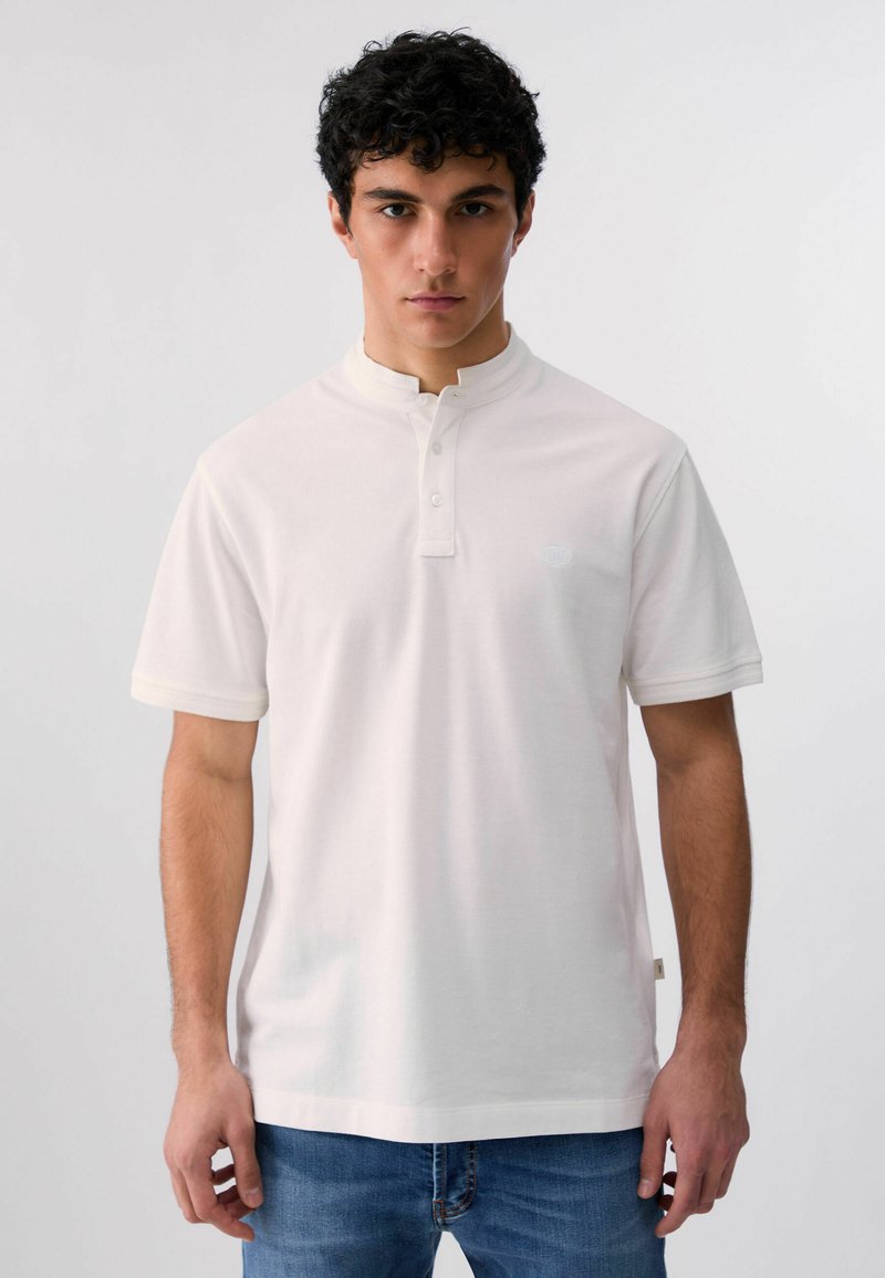 Jeune homme aux cheveux bouclés et foncés, portant un t-shirt henley blanc à manches courtes et un jean bleu, debout contre un fond clair uni.