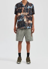 Kurzarm-Hemd mit Knöpfen im mehrfarbigen Batikmuster in Blau, Orange und Schwarz. Kombiniert mit grauen Cargoshorts mit Seitentaschen.