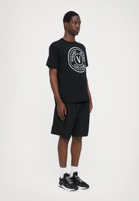 Svart bomull T-skjorte med hvit sirkulær logo, matchende svarte shorts og svarte sneakers med hvite detaljer og tekst. Enkelt, uformelt antrekk.