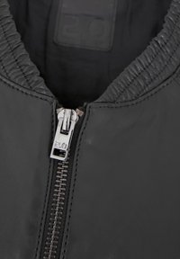 Schwarze Lederjacke mit geripptem Kragen, ausgestattet mit einem metallischen Reißverschluss in der Mitte. Die glatte Textur und das minimalistische Design werden hervorgehoben.