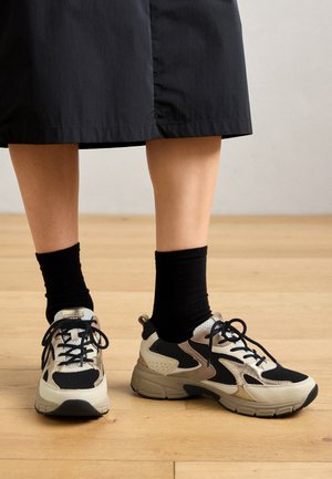 Schwarze und beige sportliche Sneakers mit einem Netz-Obermaterial, Schnürsenkeln und einer strukturierten Sohle, kombiniert mit schwarzen Socken, auf einem Holzboden.