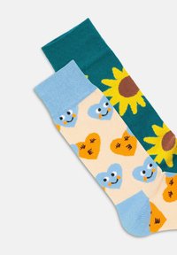 Farverige sokker: én grøn med en solblomst, den anden i beige med blå hjerter og smilende ansigt, lavet af et blødt, strækbart materiale.