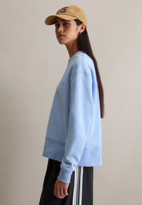 Blauwe sweatshirt met geribbelde manchetten en tailleband, met een ontspannen pasvorm. Gecombineerd met donkere broek en een beige pet.