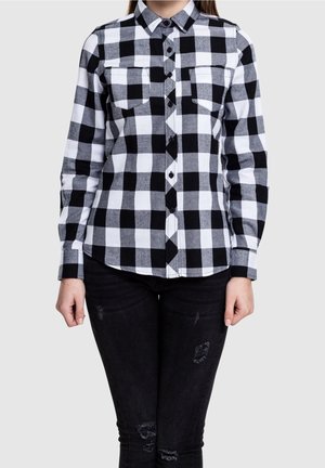 Osoba ubrana w czarno-białą koszulę w kratę zapinaną na guziki z dwoma kieszeniami na piersi, w połączeniu z czarnymi, podartymi obcisłymi dżinsami.