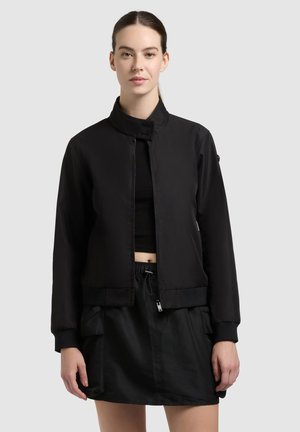 Joven con cabello oscuro recogido, que lleva una chaqueta negra con cremallera sobre un top corto negro y una falda negra con bolsillos.