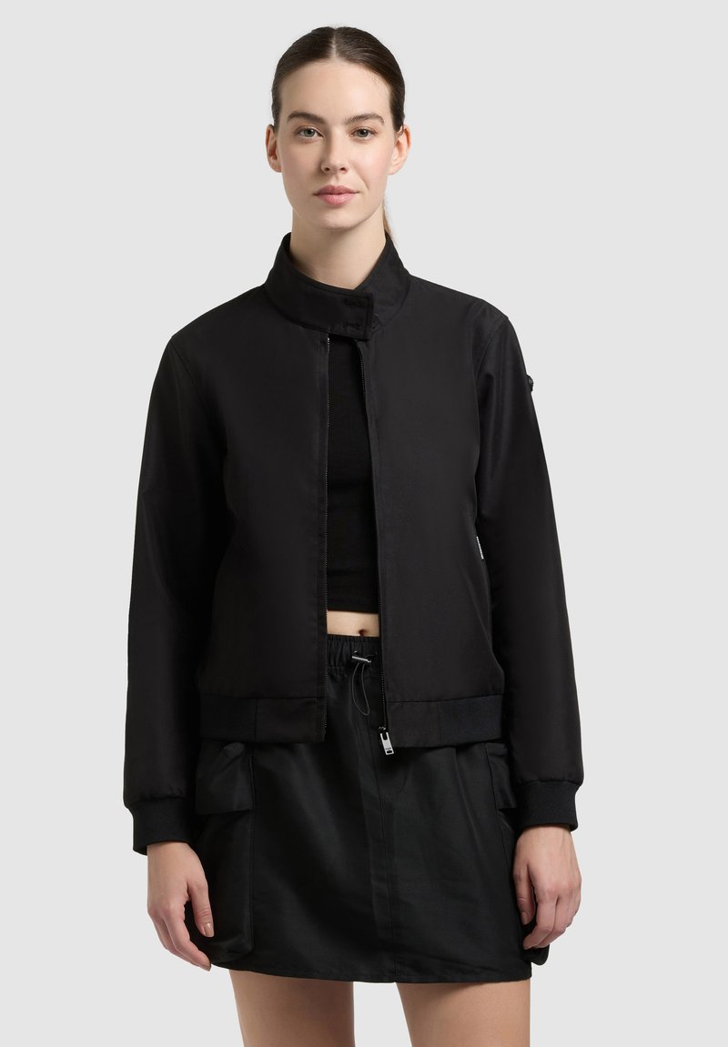 Jeune femme aux cheveux foncés tirés en arrière, portant une veste noire à fermeture éclair sur un crop top noir et une jupe noire avec des poches.