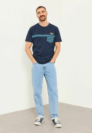 Mann mit Schnurrbart, der ein marineblaues T-Shirt mit Grafik, hellblaue Jeans und marineblaue Sneakers trägt, steht entspannt an einer weißen Wand.