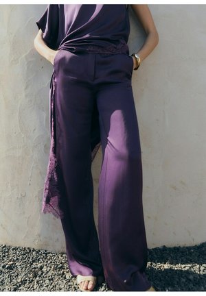 Persona che indossa pantaloni larghi di raso viola e top abbinato con dettagli in pizzo, in piedi contro una parete beige strutturata su ghiaia.