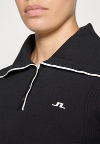 Black polo shirt with a textured surface, featuring a white collar trim, three buttons, and a small white logo on the chest.