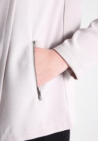 Poche gris clair zippée sur un tissu texturé, avec une main placée à l'intérieur. La poche est équipée d'une fermeture éclair en métal argenté pour une fermeture sécurisée.