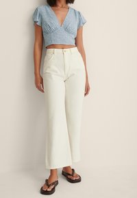 Crop top bleu clair style cache-cœur avec manches volantées, associé à un jean blanc taille haute à jambes larges et des sandales tongs foncées.