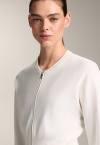 Bela zip-up jopica z zaobljenim ovratnikom, gladko teksturo in prilegajočim dizajnom. Ima srebrno zadrgo in diskretne šivane detajle.