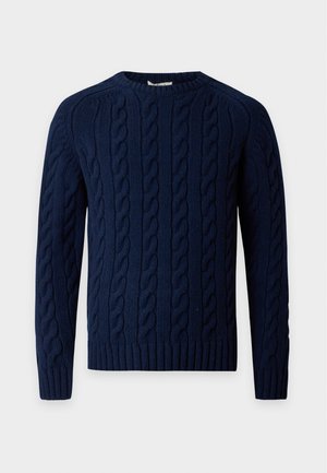Homme portant un pull en maille torsadée bleu marine, un jean bleu délavé ample et des chaussures noires, debout les mains dans les poches devant un fond blanc uni.