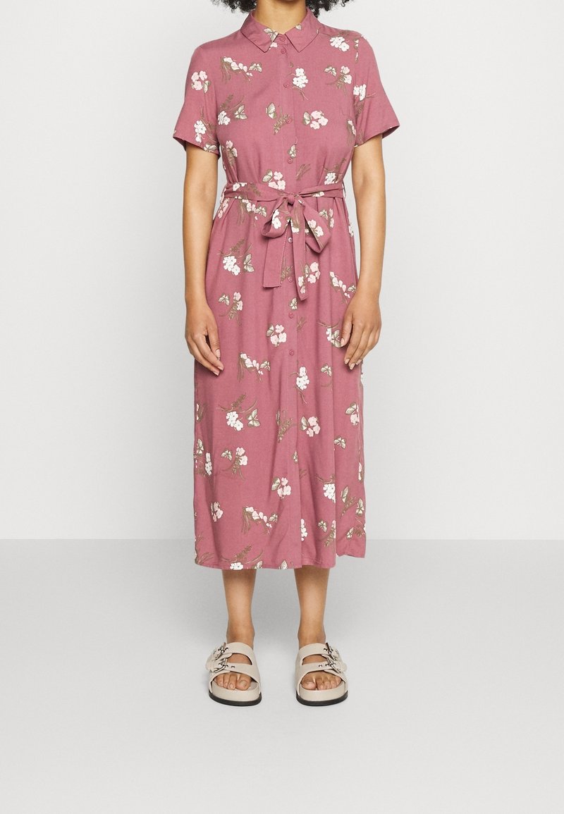 Femme portant une robe midi fleurie rose à manches courtes, avec un col, une ceinture à la taille et des sandales beiges à boucles.