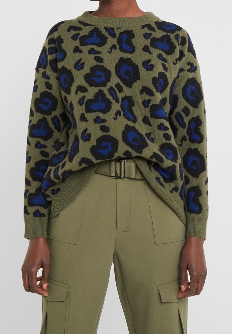 Pull vert olive avec imprimé léopard bleu et noir, associé à un pantalon cargo vert olive avec poches avant et ceinture.
