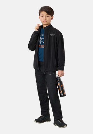 Chłopiec ubrany w czarną kurtkę zapinaną na suwak i spodnie, trzyma czarną butelkę na wodę z pomarańczowymi nadrukami zwierząt, stoi na jednolitym tle.