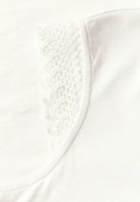 Primer plano de una tela blanca con un inserto de encaje texturizado cerca del escote. Textura suave con un diseño de cuello redondeado.