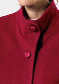 Roter Wollmantel mit hohem Kragen und zwei auffälligen runden Knöpfen. Die weiche Textur und subtile Stickdetails verbessern sein Design.