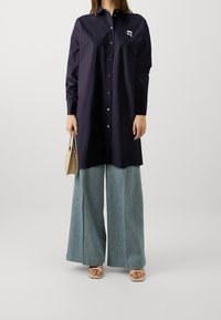 Dunkelblaue Hemdbluse mit langen Ärmeln und kleinem Logo, kombiniert mit weiten hellblauen Jeans und beigen Sandalen, während eine beige Handtasche gehalten wird.