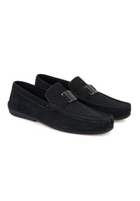 Schwarze Wildleder-Loafer mit flacher Sohle und einem dekorativen silbernen Schnallen-Detail an der Vorderseite. Die Nähte sind entlang der Kanten sichtbar.