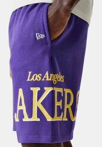 Pantaloni corti viola con grande scritta gialla "LAKERS" e "Los Angeles" sopra. Realizzati in tessuto morbido, con tasca laterale e dettaglio del logo.