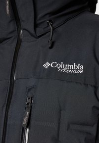 Crna Columbia Titanium jakna s glatkom teksturom, sa zip zatvorenim džepom i podesivom kapuljačkom. Logo vezan na prsima.