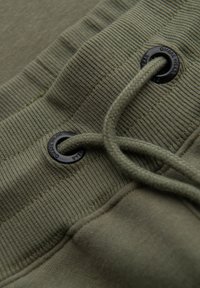 Nærbillede af olivengrøn ribbet linning med flettet snor trukket gennem sorte metaløjne på stofbeklædning.