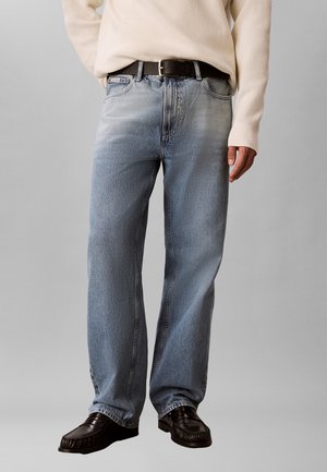 Man wearing loose light blue jeans with a black belt, cream sweater, and black leather loafers, standing against a plain background.