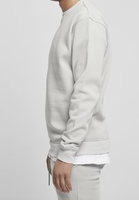 Felpa grigia chiara con maniche lunghe, polsini e fondo a costine, scollatura rotonda, texture liscia e stile a strati sopra una maglietta bianca.