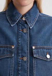 Donkerblauwe denimjack met een klassieke kraag, zilveren knopen en twee voorzakken. Beschikt over contrasterende stiksels en een gestructureerde afwerking.