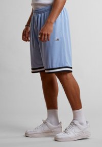 Shorts de sport bleu clair avec un ourlet rayé noir et blanc, arborant un petit logo sur le côté. Portés avec des baskets blanches et des chaussettes.