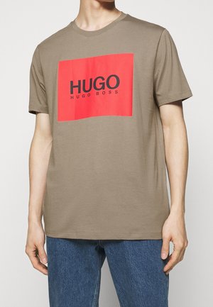 Osoba ubrana w brązową koszulkę z krótkim rękawem z czerwonym prostokątem i czarnym logo "HUGO HUGO BOSS" na klatce piersiowej oraz niebieskie dżinsy.
