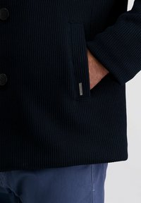 Marineblau gerippter Strickmantel mit vier Knöpfen, schrägen Seitentaschen und einem Metall-Logo-Detail. Verfügt über einen strukturierten Stoff und eine definierte Form.