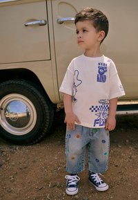Witte t-shirt met blauwe grafische ontwerpen, inclusief tekst en patronen, gecombineerd met lichtblauwe denim shorts met kleurrijke glimlach- patches en donkere sneakers.