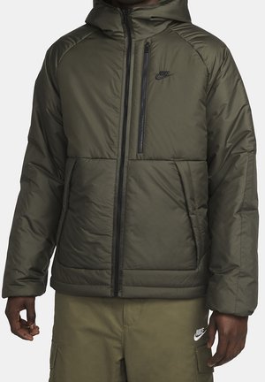 Homme portant une doudoune Nike vert olive avec capuche et fermeture éclair noire, associée à un pantalon cargo Nike assorti.