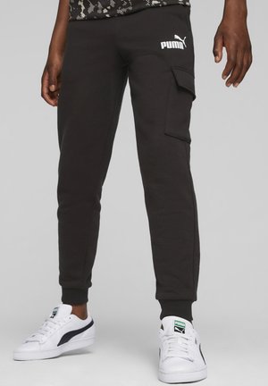Pantalons de survêtement noirs avec poche cargo, coupe fuselée, poignets côtelés et logo Puma blanc. Portés avec des baskets blanches présentant des accents verts.