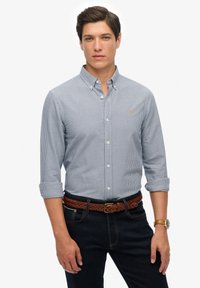Camisa de cuadros en azul claro y blanco con mangas remangadas, combinada con jeans oscuros y un cinturón de cuero trenzado marrón.