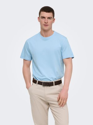 Man with short brown hair wearing light blue t-shirt, beige pants, and dark belt, standing with hands in and near pockets on white background.